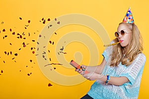 Portrait of funny blond woman in birthday hat and red confetti on yellow background. Celebration and party.