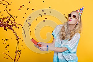 Portrait of funny blond woman in birthday hat and red confetti on yellow background. Celebration and party.