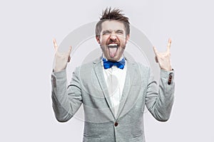 Portrait of funny crazy handsome bearded man in casual grey suit and blue bow tie standing with rock gesture, closed eyes and