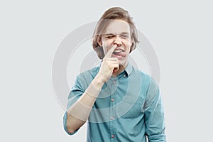 Portrait of funny crazy handsome long haired blonde young man in blue casual shirt standing drilling his nose and tongue out