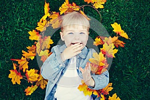 Portrait of funny cute smiling white Caucasian toddler child girl with blond hair lying on green grass with yellow autumn leaves