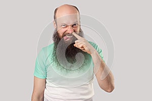 Portrait of funny dirty middle aged bald man with long beard in light green t-shirt standing drilling with finger on his nose,