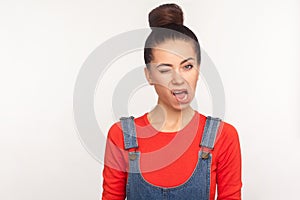 Portrait of funny displeased girl with hair bun in denim overalls winking to camera, looking with ugly skewed face