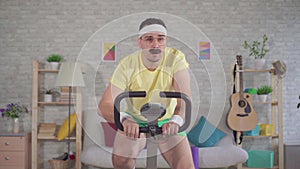 Portrait funny energetic man athlete from the 80`s with a mustache engaged at home on a stationary bike