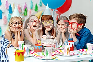 Portrait of funny group of children wear party caps, big spectacles, look with big appetite on birthday cake, want to