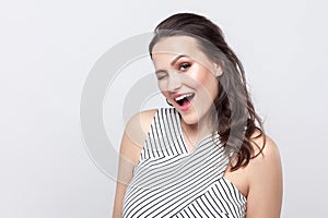 Portrait of funny happy beautiful young brunette woman with makeup and striped dress standing, winking and looking at camera with