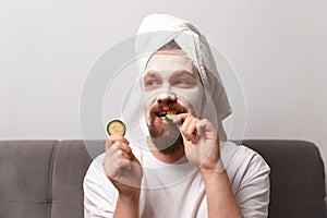 Portrait of funny happy man in white tshirt applying cucumber slices over clay mask on his face. Self care morning spa