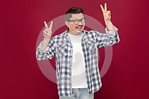 Portrait of funny happy middle aged man in casual checkered shirt, eyeglasses standing with peace or victory sign and looking at