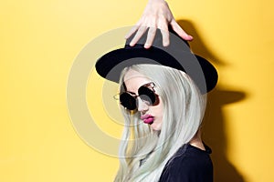 Portrait of funny hipster girl, touching her hat with hand, wearing sunglasses, isolated on yellow background.