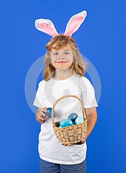 Portrait of funny kid with bunny ears hold easter basket with easter eggs, isolated on blue studio background. Easter