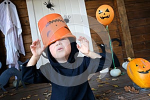 Portrait of funny little kid in wizard costume near door of house, putting candy bucket for trick or treat on head and saing boo.