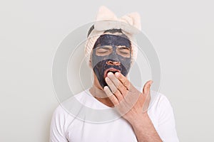 Portrait of funny man wearing white t shirt and female`s hair band standing with mud mask and yawning, covering mouth with palm,