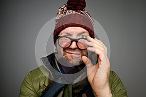 Portrait of a funny mature man in warm winter clothes looking at camera through glasses over gray background.