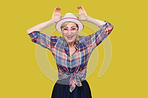 Portrait of funny modern stylish mature woman in casual style with hat and eyeglasses standing with bunny ears gesture and looking