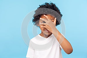 Portrait of funny nosy adorable little boy with curly hair covering face with hand and looking through fingers