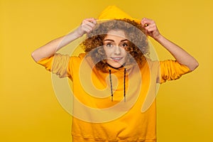 Portrait of funny positive girl with curly hair wearing urban style hoodie, putting on hood and smiling