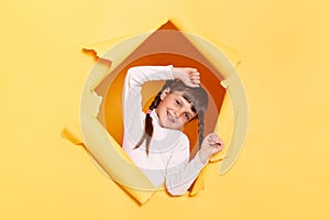 Portrait of funny positive little girl with braids wearing casual shirt looking through torn hole in yellow paper, keeps hands