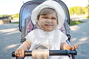 Portrait of funny smiling baby girl in white hat sitting in stroller, make faces