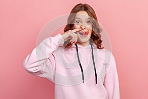 Portrait of funny teen curly haired girl in hoodie drilling nose fooling around, showing tongue out and picking nose with comical