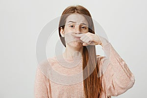 Portrait of funny upbeat ginger girl holding index finger above nose as if it is moustache and lifting one eyebrow