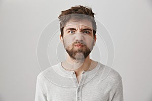 Portrait of funny weird guy with messy hair and beard making faces, puckering eyebrows and sulking, standing over gray