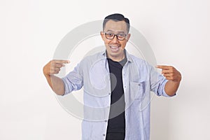 Portrait of funny young Asian man smiling and pointing to presenting something in front his body. Isolated on white background