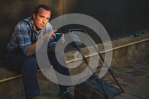 Portrait of a gardener man looking at camera, sitting near a lawn mower with cup of coffee in hands, relaxing at sunset