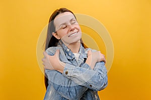 Portrait of gentle lovely beautiful young woman standing with closed eyes hugging herself, being selfish, wearing denim jacket,