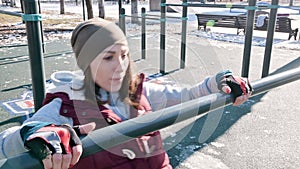 Portrait of girl athlete in a hat with gloves and a warm vest doing exercises on uneven bars at an outdoor sports field