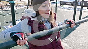 Portrait of girl athlete in a hat with gloves and a warm vest doing exercises on uneven bars at an outdoor sports field