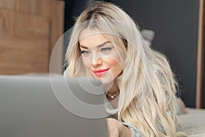 Portrait of a girl behind a laptop screen. Beautiful blonde woman lying on the bed and looks at the laptop screen