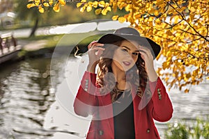 Portrait of a girl in black hat with a river on a background reflecting the sun`s rays and yellow autumn trees