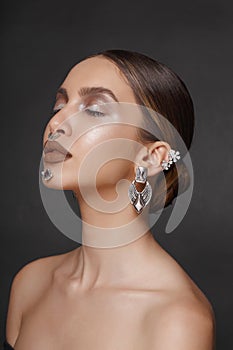 Portrait of a girl with closed eyes, makeup and jewelry applied to the face, posing with attitude, over gray background.
