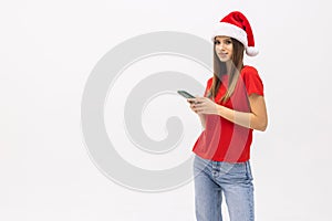 Portrait of a girl dressed in red christmas hat holding mobile phone while standing and looking at camera isolated over white