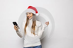 Portrait of a girl dressed in red christmas hat holding mobile phone while standing and looking at camera isolated over white