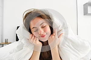 Portrait of girl feeling sleepy after waking up early, lying in bed under blanket and smiling