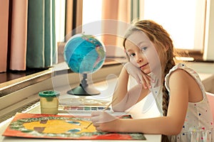 Portrait of girl kid is painting with colors gouache and brush picture while sitting at table on home room background.