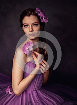 Portrait of a girl in a lilac lush dress with bare shoulders, she holds branches of lilac orchid flowers in her hands.