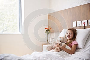 Portrait Of Girl Lying In Bed In Hospital Ward Hugging Teddy Bear