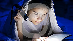 Portrait of girl lying under blanket at night and reading book with light torch