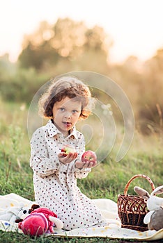 Portrait of a girl on a meadow in the sunset sunlight. Little girl in a light overalls sits on a blanket next to basket with harve