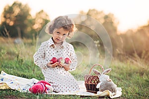 Portrait of a girl on a meadow in the sunset sunlight. Little girl in a light overalls sits on a blanket next to basket with harve