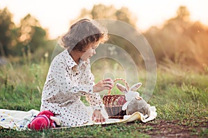 Portrait of a girl on a meadow in the sunset sunlight. Little girl in a light overalls sits on a blanket next to basket with harve