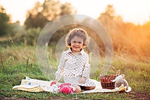 Portrait of a girl on a meadow in the sunset sunlight. Little girl in a light overalls sits on a blanket next to basket with harve