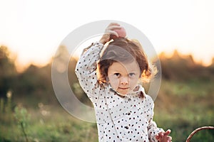 Portrait of a girl on a meadow in the sunset sunlight. Little girl in a light overalls sits on a blanket next to basket with harve