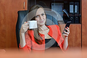 Portrait of a girl in a red business suit sitting resting in a chair, drinking tea and using a smartphone. break time, social netw