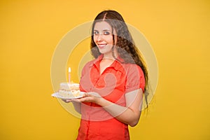 Portrait of a girl in a red shirt with a piece of cake and candle on a white plate in her hands isolated on a yellow background