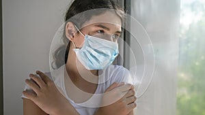 Portrait of girl sick corona virus. little teenager girl in a medical mask is sad at the window hugs herself over the