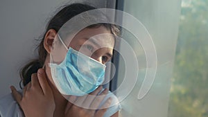 Portrait of girl sick corona virus. little teenager girl in a medical mask is sad at the window hugs herself over the