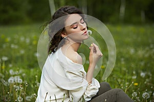 Portrait of a girl sitting in a field on the spring grass among dandelion flowers. Cheerful girl enjoys Sunny spring weather.